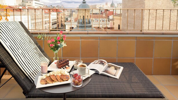 A balcony with a wicker lounge chair, a tray with pastries, a glass of juice, a cup of coffee, a vase with flowers, and an open magazine. - Hotel Francisco I (Madrid)