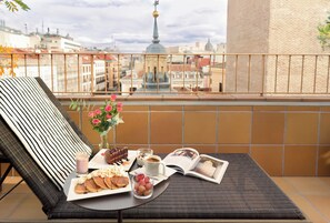 Un balcón con una silla lounge de mimbre, una bandeja con pasteles, un vaso de jugo, una taza de café, un florero con flores y una revista abierta.