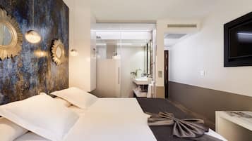 Una habitación de hotel moderna con una cama grande, un espejo con marco dorado, un televisor de pantalla plana y un baño visible al fondo.