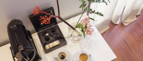 A small table with a coffee maker, two cups of coffee, a vase with flowers, and a tray with a box and a remote control.