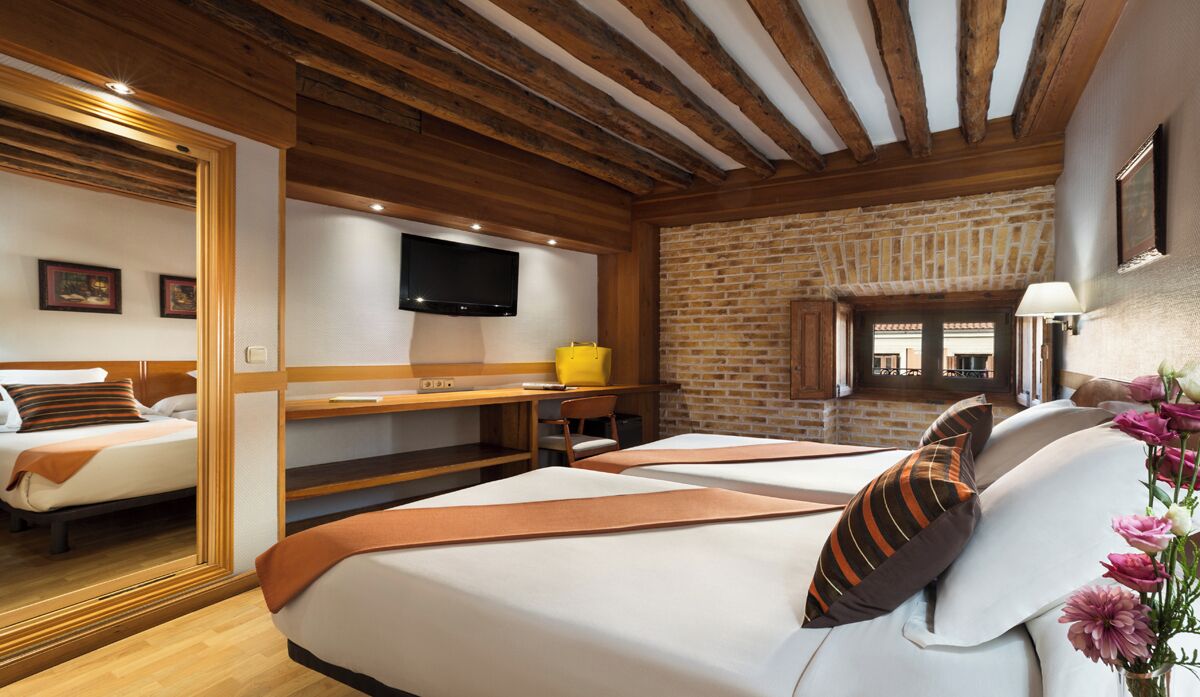 A hotel room with wooden beams, a brick wall, a TV, a bed, a desk, and a chair.