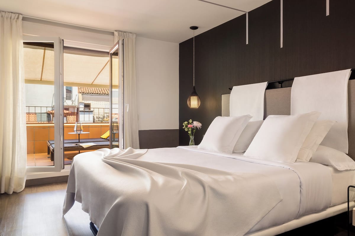 A modern hotel room with a large bed, a glass table, a nightstand, and a view of the outdoor balcony.