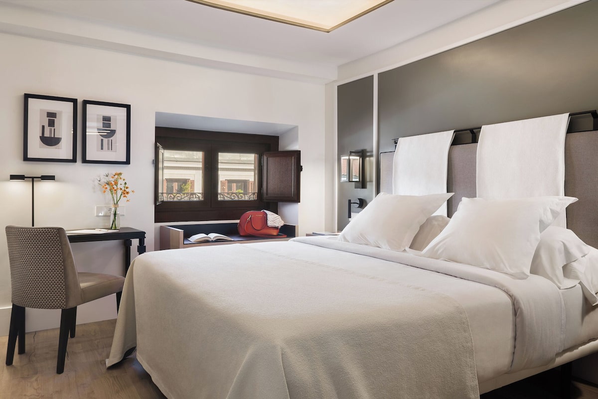 A modern hotel room with a large bed, a desk, and a chair. There is a window with a view of buildings outside.