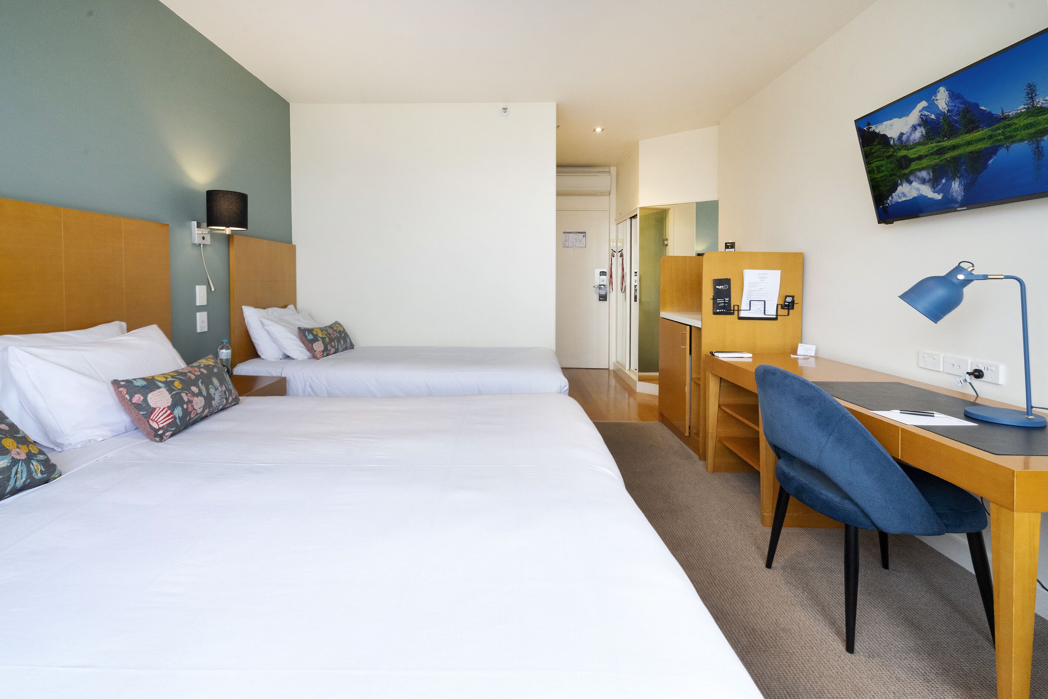 A hotel room with two beds, a desk, a chair, a desk lamp, and a wall-mounted TV.