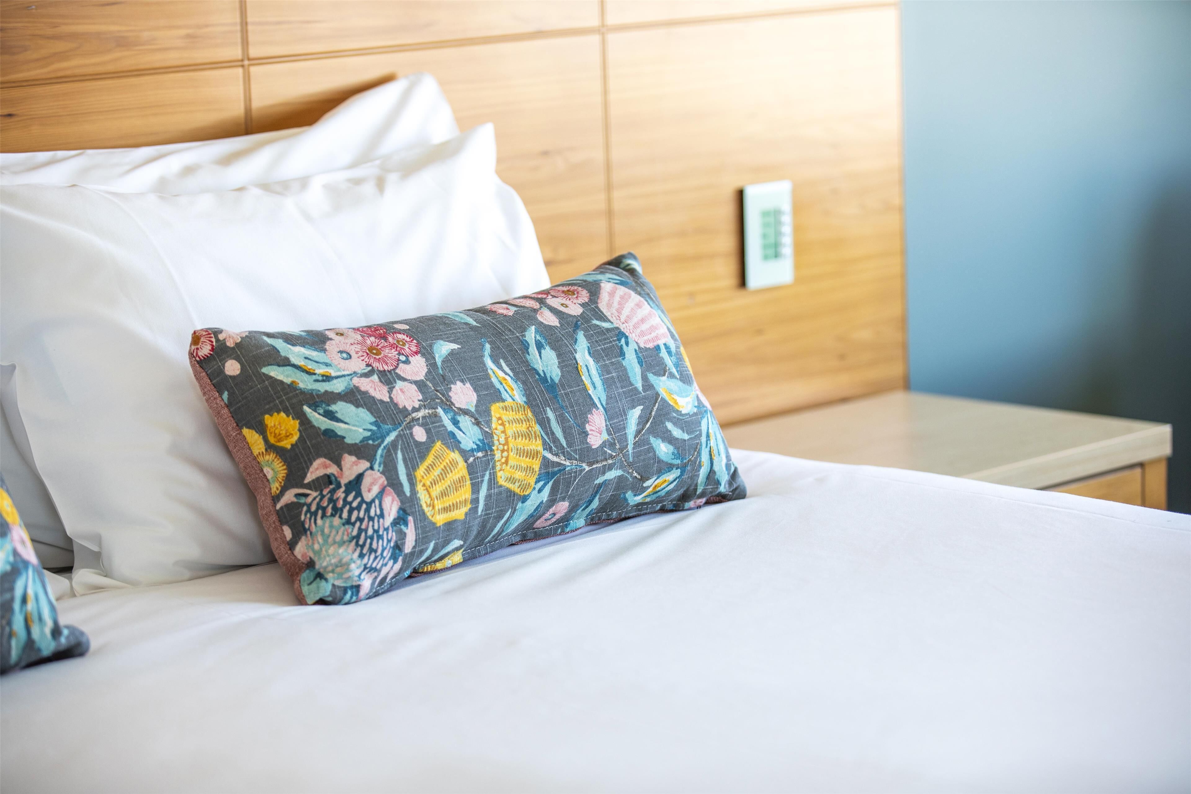 A neatly made bed with white and floral-patterned pillows, a wooden headboard, and a small bedside table.