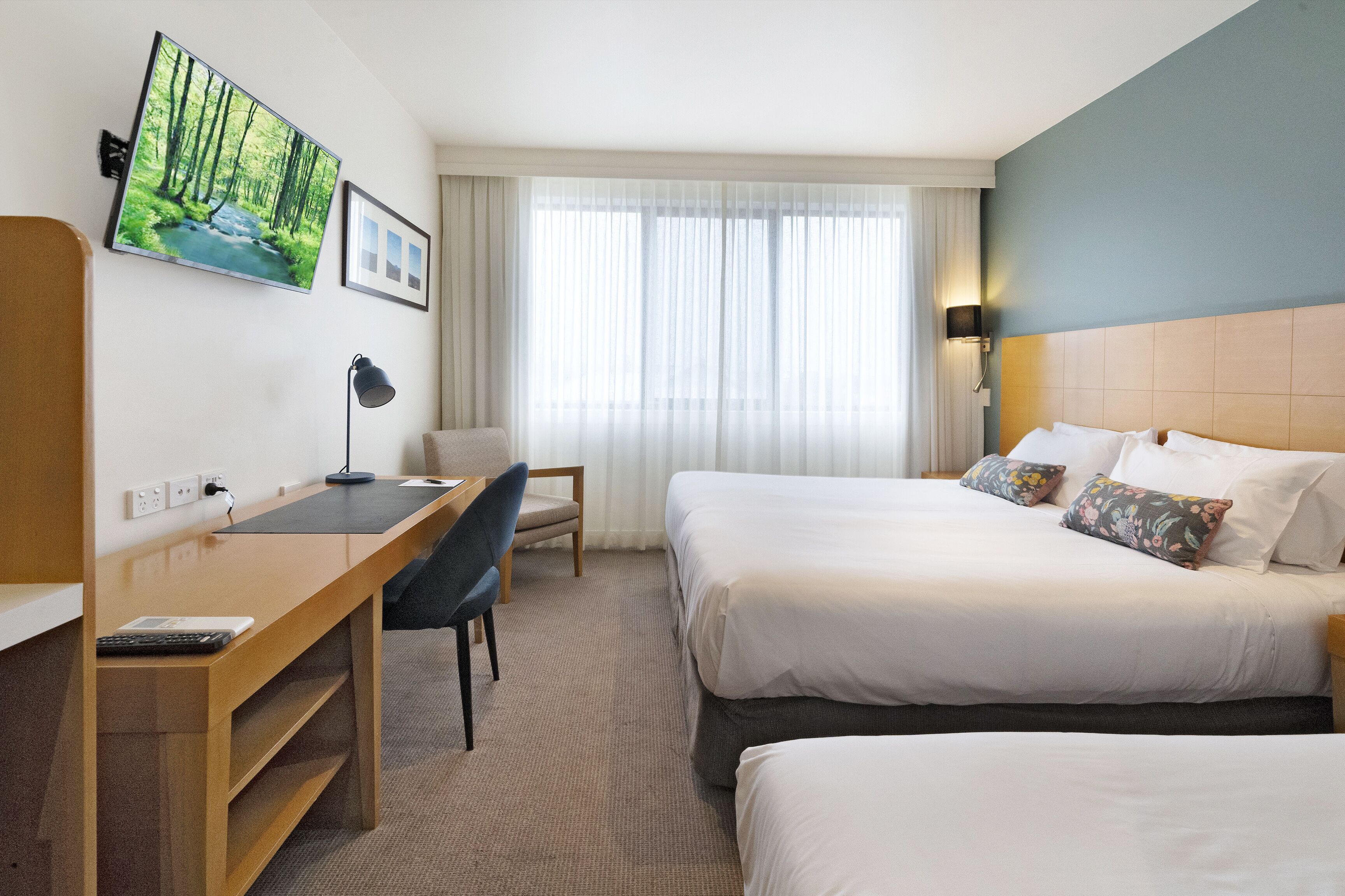 Habitación de hotel con dos camas, un escritorio, una silla, un televisor y una ventana con cortinas translúcidas.