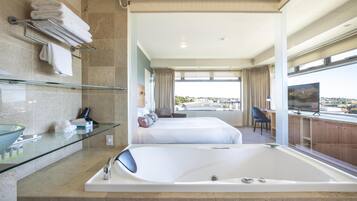 Quarto de hotel moderno com box de vidro, uma banheira grande, cama com vista para a cidade e televisão em um móvel de madeira.