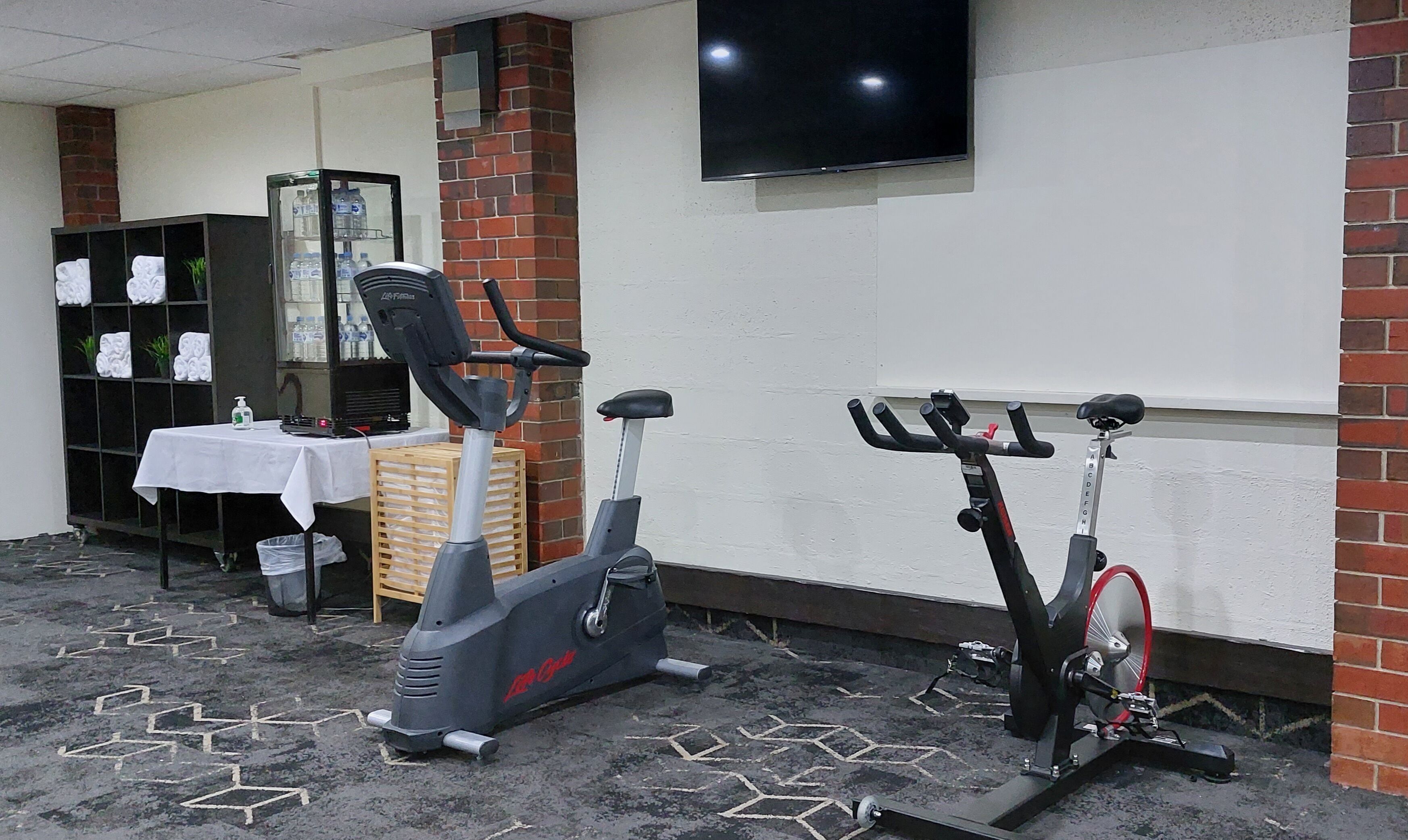 Quarto com duas bicicletas ergométricas, uma mesa com toalha branca, uma estante com toalhas e uma televisão fixada na parede.