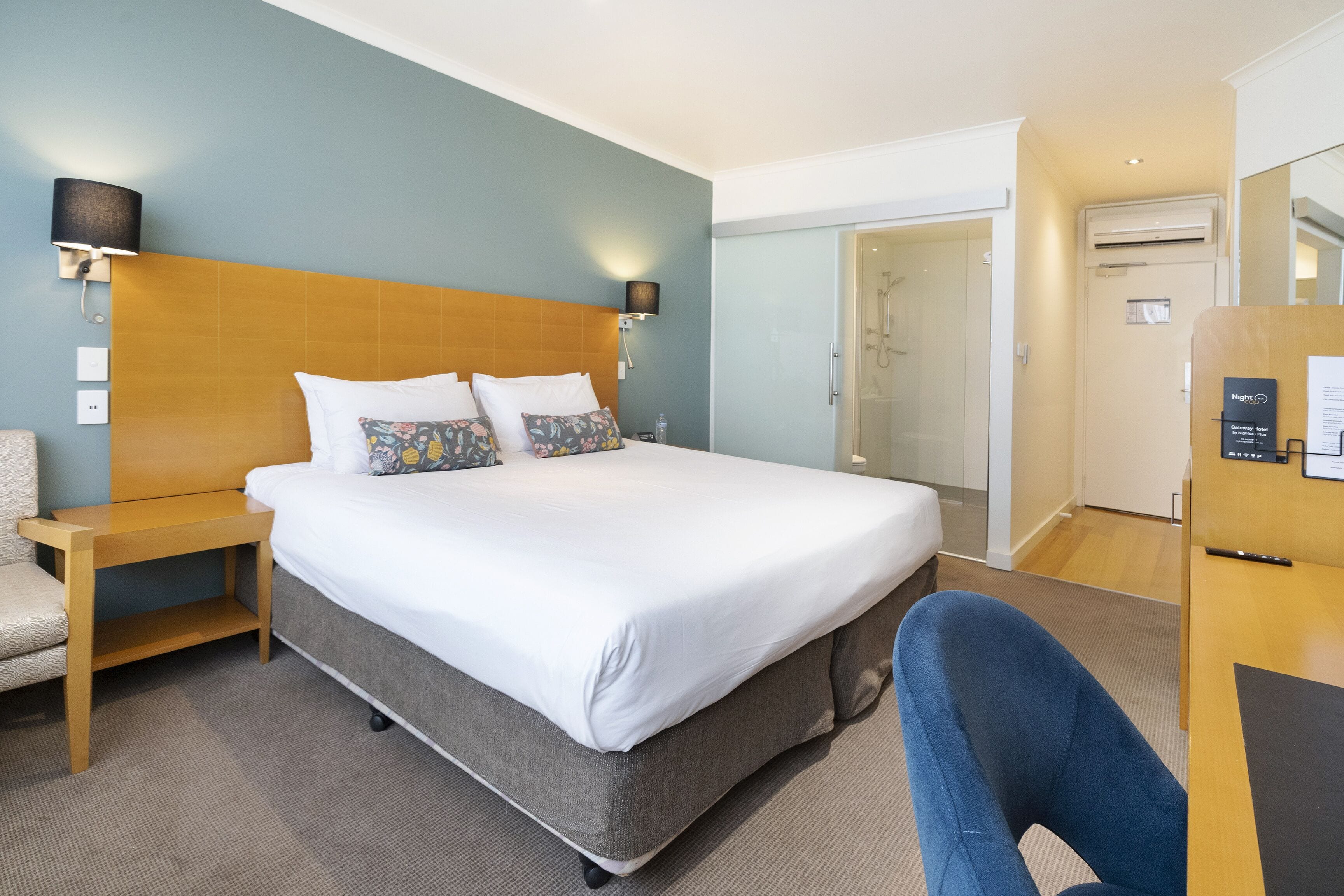 A hotel room with a large bed, a bedside table, a chair, a desk, and a shower area.