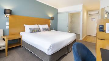 A hotel room with a large bed, a bedside table, a chair, a desk, and a shower area.