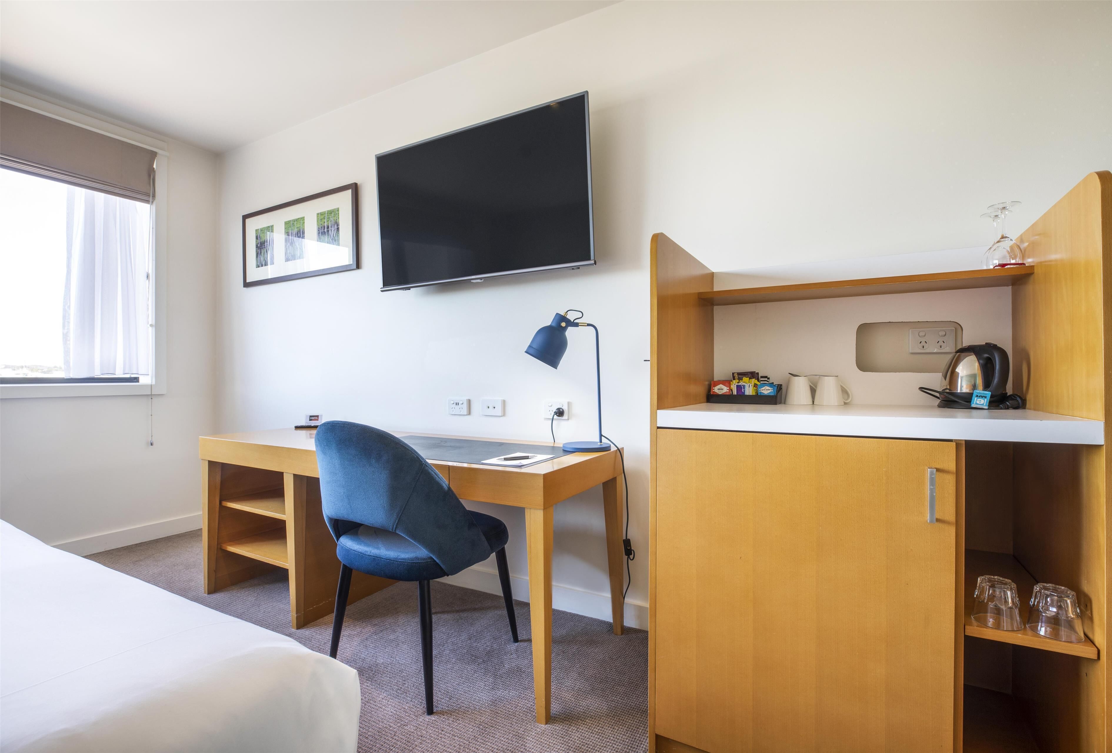 A hotel room with a bed, a desk with a chair, a TV, and a kitchenette area.