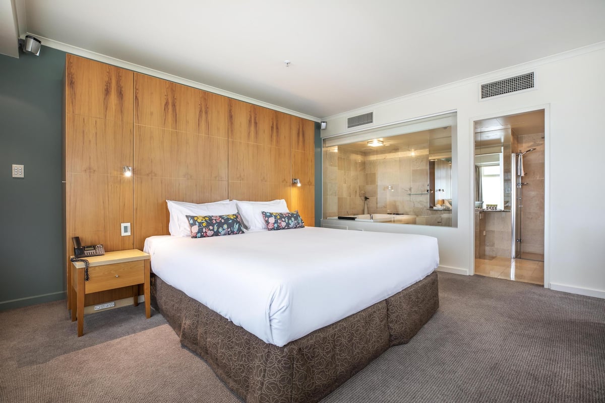 Habitación de hotel con una cama grande, un panel de pared de madera, una mesita de noche con teléfono y un baño con ducha de vidrio.
