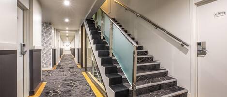 Un pasillo de hotel con piso alfombrado, una escalera con barandilla de vidrio y una puerta con letrero de número.