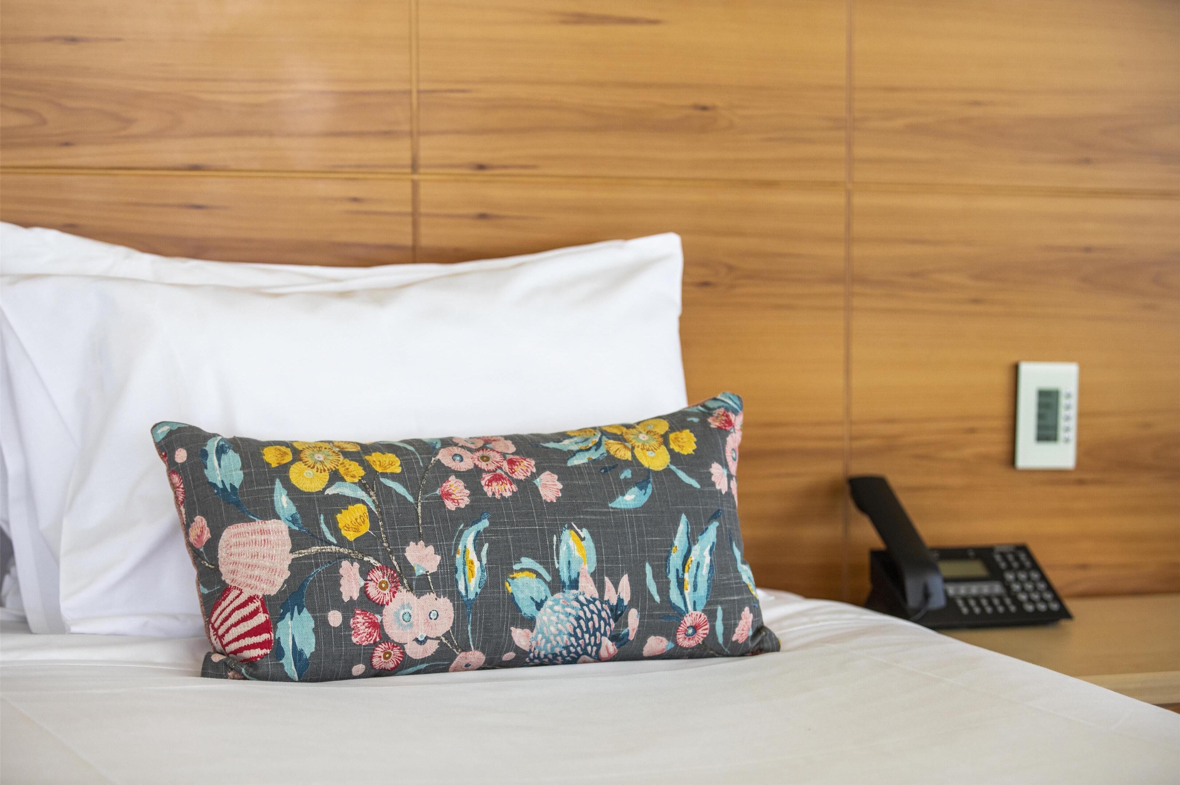 Una cama con sábanas blancas, un cojín estampado con motivos florales y un teléfono sobre una pared de madera.