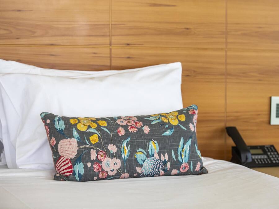 Una cama con sábanas blancas, un cojín estampado con motivos florales y un teléfono sobre una pared de madera.