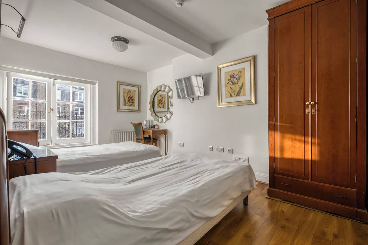 A hotel room with two beds, a wooden wardrobe, a desk with a chair, a radiator, and framed pictures on the wall.