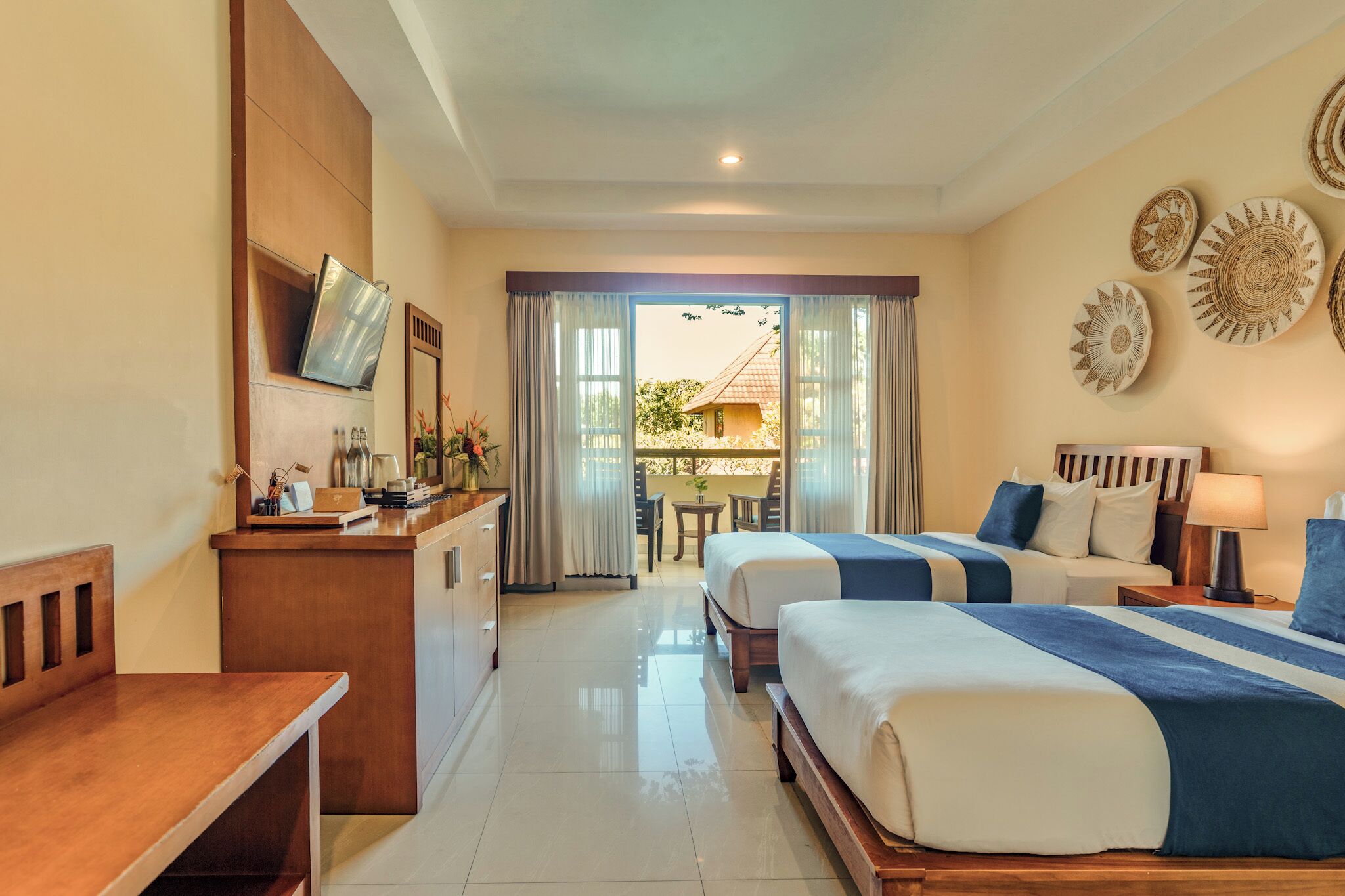 Habitación de hotel con dos camas, un escritorio, un televisor y un balcón con vista a una casa.