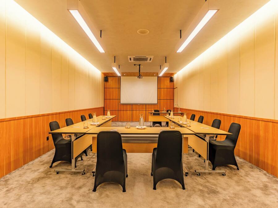 Een vergaderruimte met een lange tafel, zwarte stoelen, een projectiescherm en houten paneelwanden.