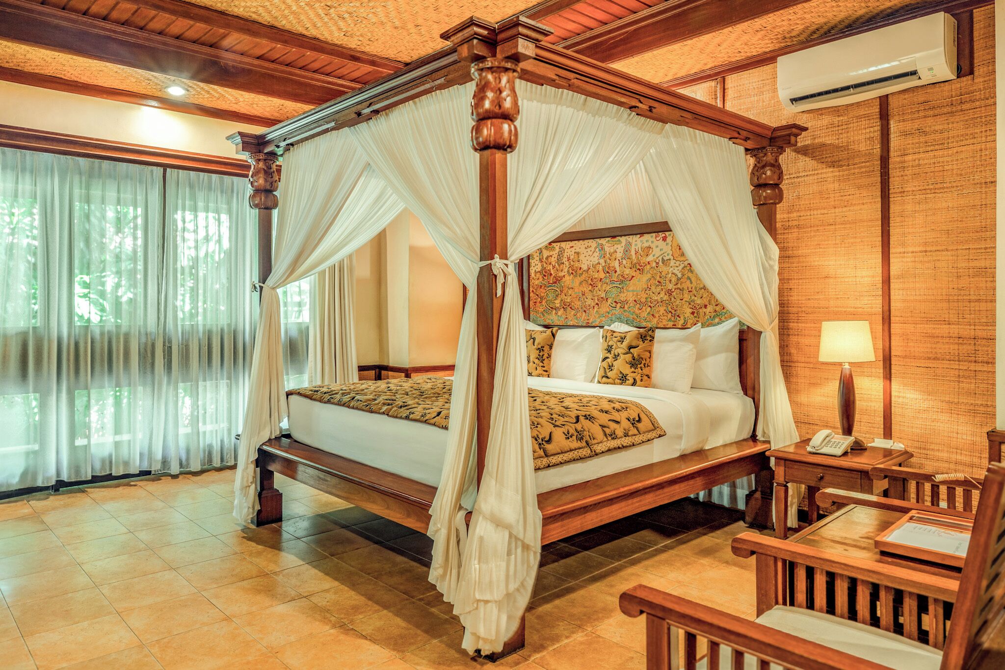 A four-poster canopy bed with a wooden frame and white drapes, a bedside table with a lamp, and a view of a garden through large windows.