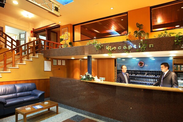 Zona de recepción de hotel con mostrador de salida, una escalera, mostrador de recepción y un área de descanso con sofá y mesa de centro.