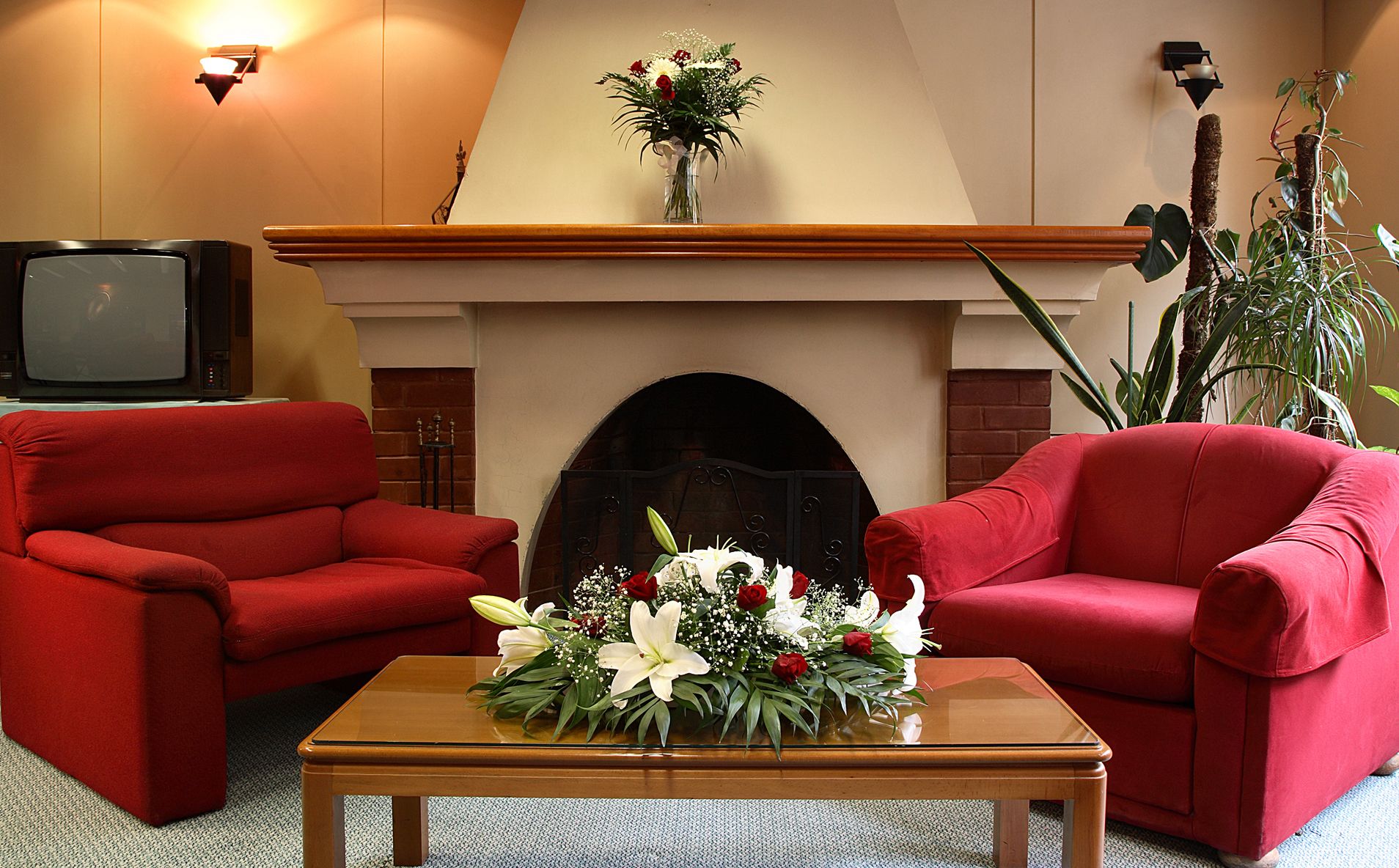 A room with a fireplace, two red armchairs, a coffee table with a floral arrangement, and a television on a stand.