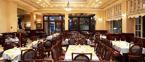 Un restaurante espacioso con múltiples mesas y sillas, un candelabro y ventanas grandes.