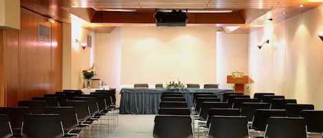 Un salón de conferencias con filas de sillas, una mesa con mantel azul y un atril con el nombre del hotel.