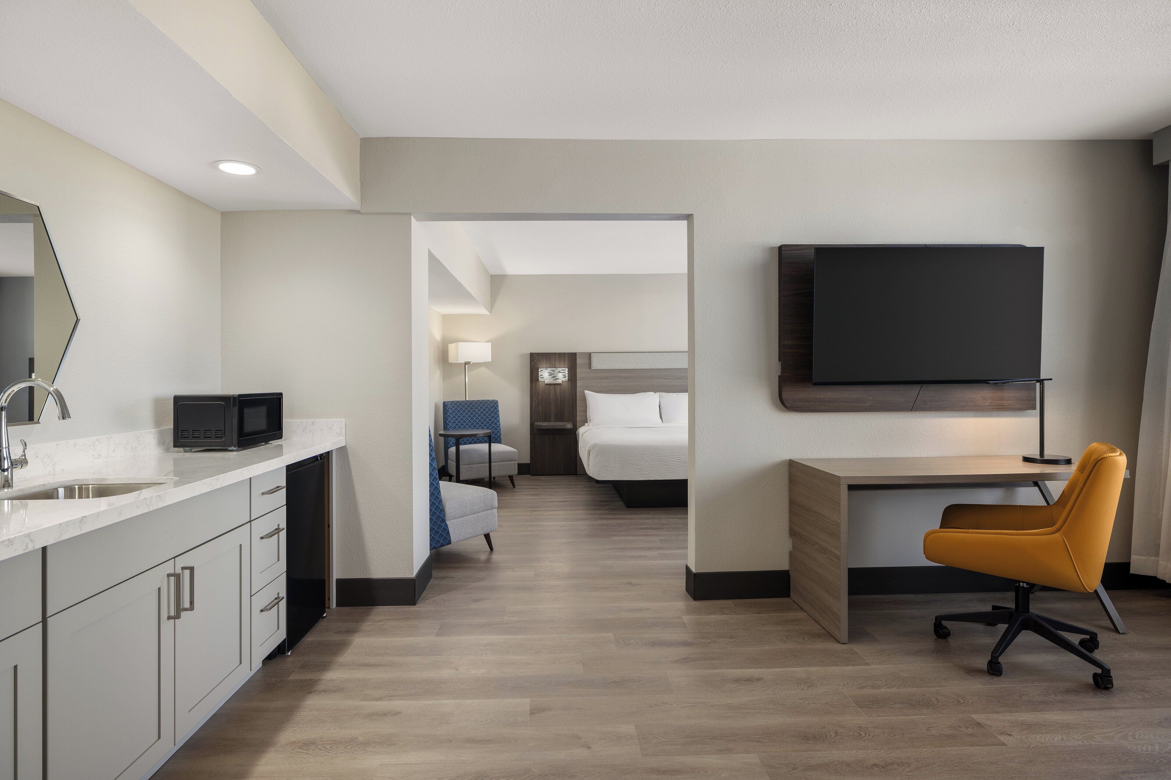 Une chambre d’hôtel moderne équipée d’une kitchenette, d’un téléviseur à écran plat fixé au mur, d’un bureau avec une chaise et d’un lit avec une tête de lit.