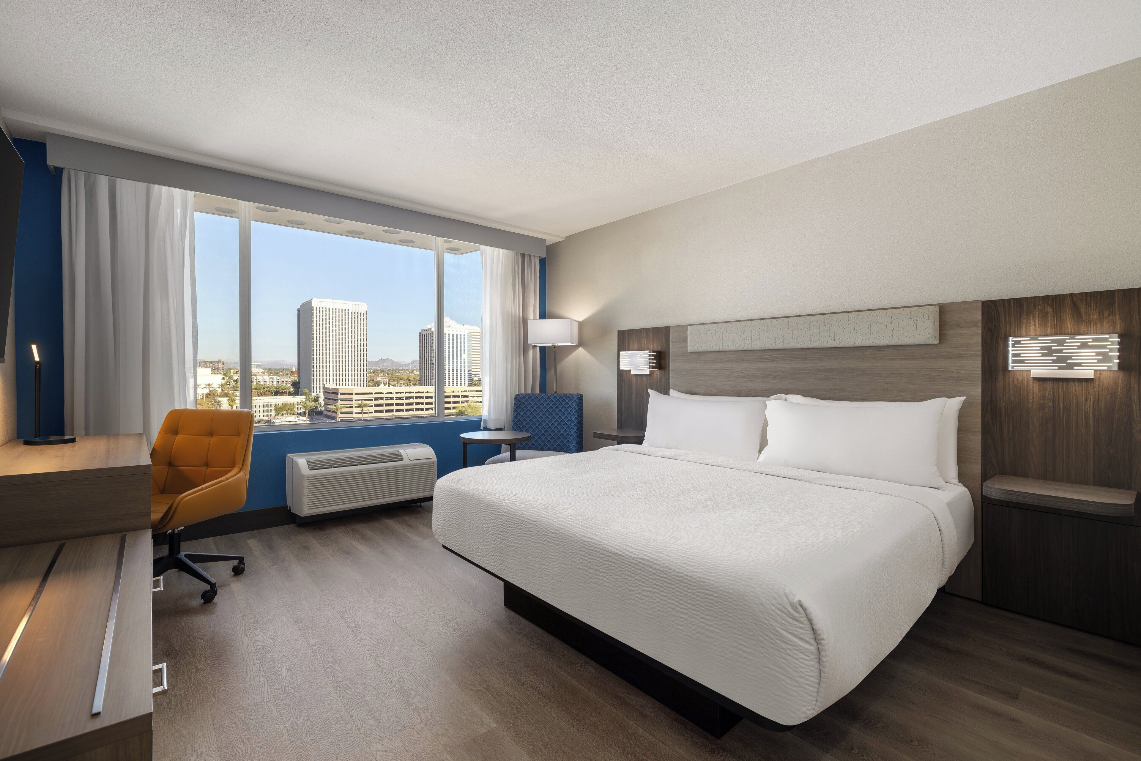 A hotel room with a bed, a desk, a chair, a window with a view of buildings, and a wall-mounted light fixture.