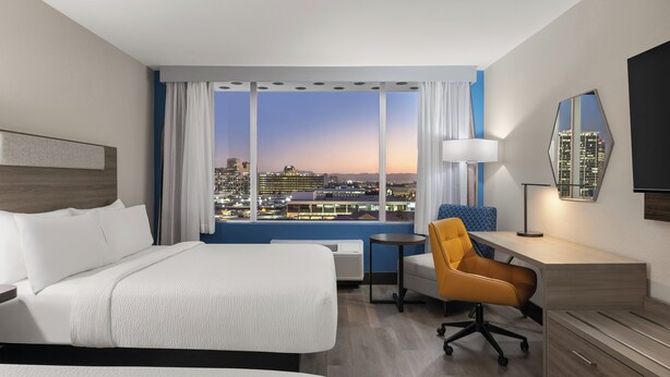 A hotel room with a bed, a desk, a chair, and a large window with a city view.