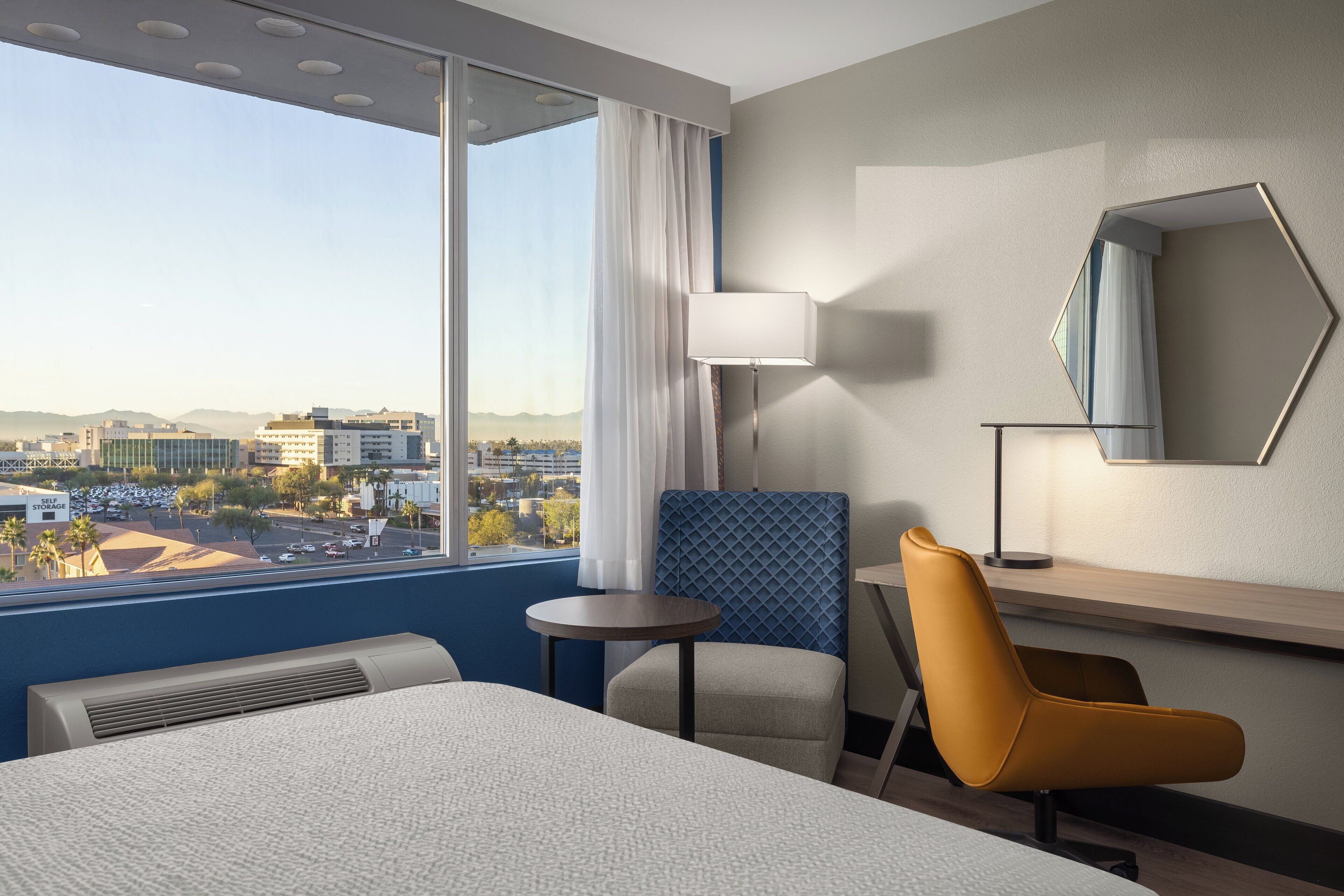 A hotel room with a large bed, a desk, a chair, a lamp, and a view of the city.