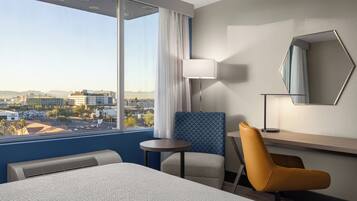 A hotel room with a large bed, a desk, a chair, a lamp, and a view of the city.
