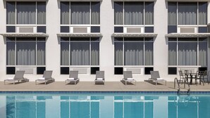 A swimming pool with lounge chairs and a sunbed area, surrounded by a building with large windows and air conditioning units.