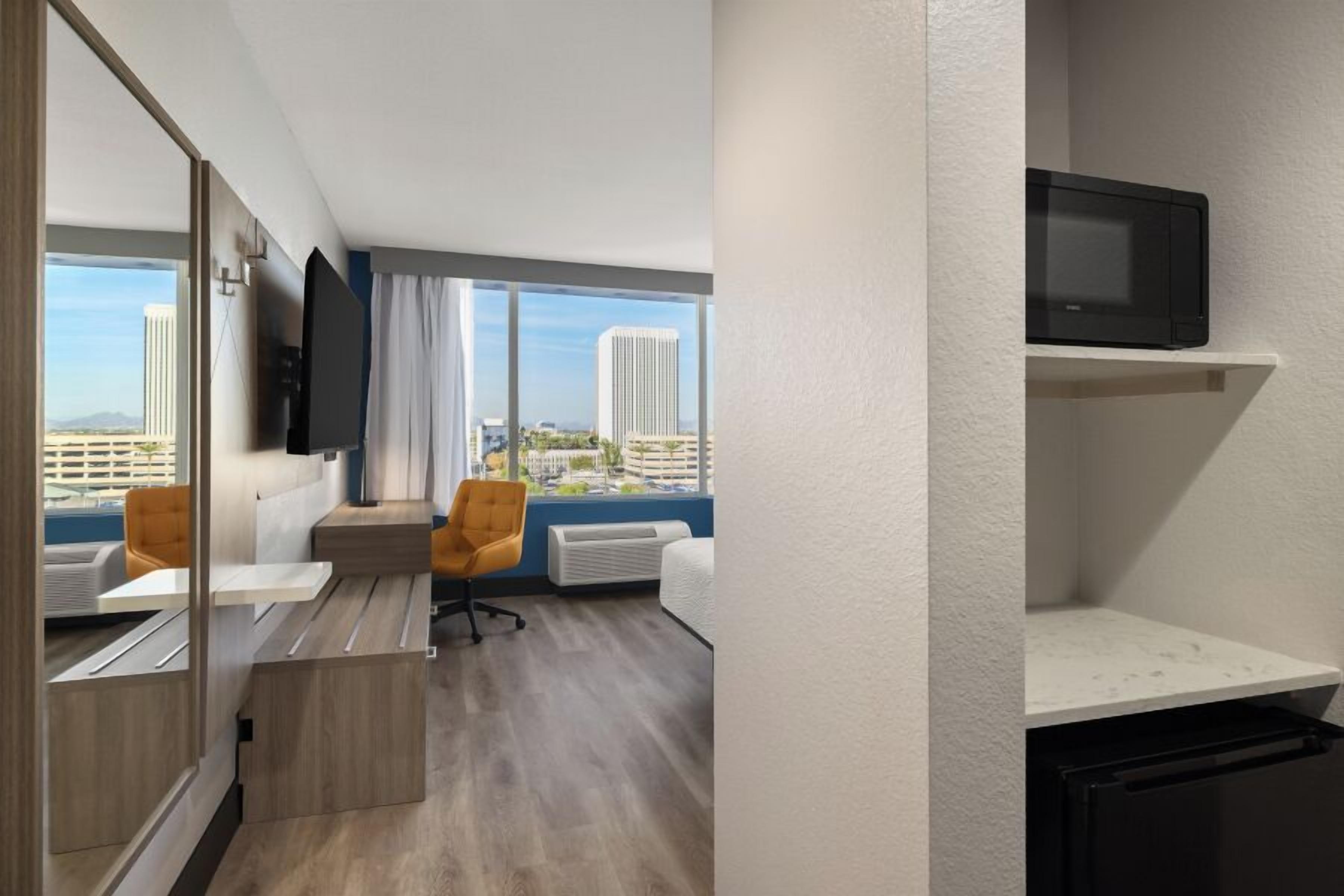 Una habitación de hotel moderna con un ventanal grande, un televisor de pantalla plana, una pequeña cocina y una zona de estar con una silla y una mesa.