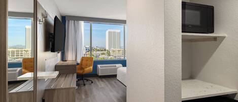 A modern hotel room with a large window, a flat-screen TV, a sofa, a chair, and a small desk.