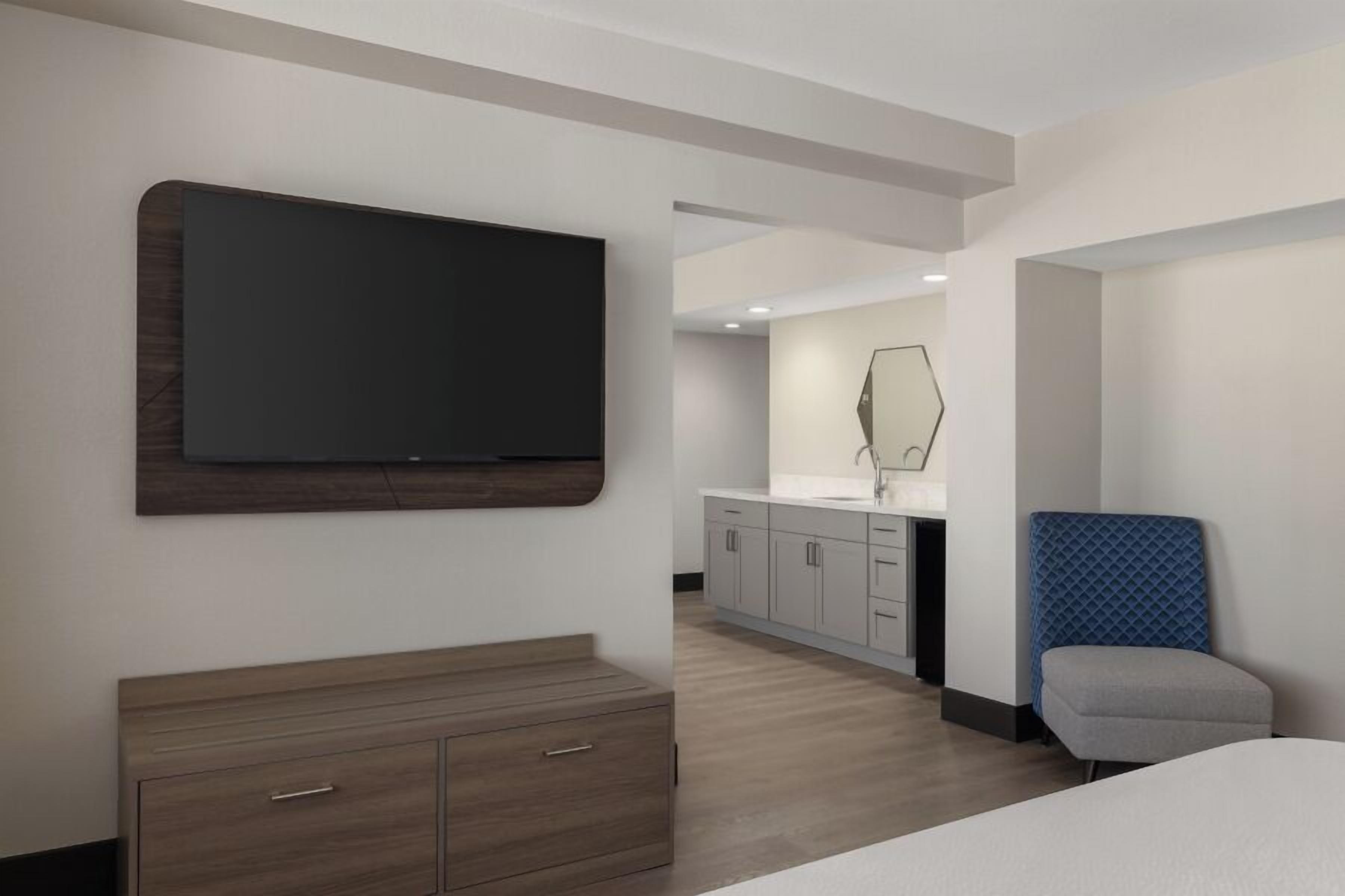 A modern hotel room with a large flat-screen TV, a wooden dresser, a blue cushioned chair, and a bathroom visible through an open door.