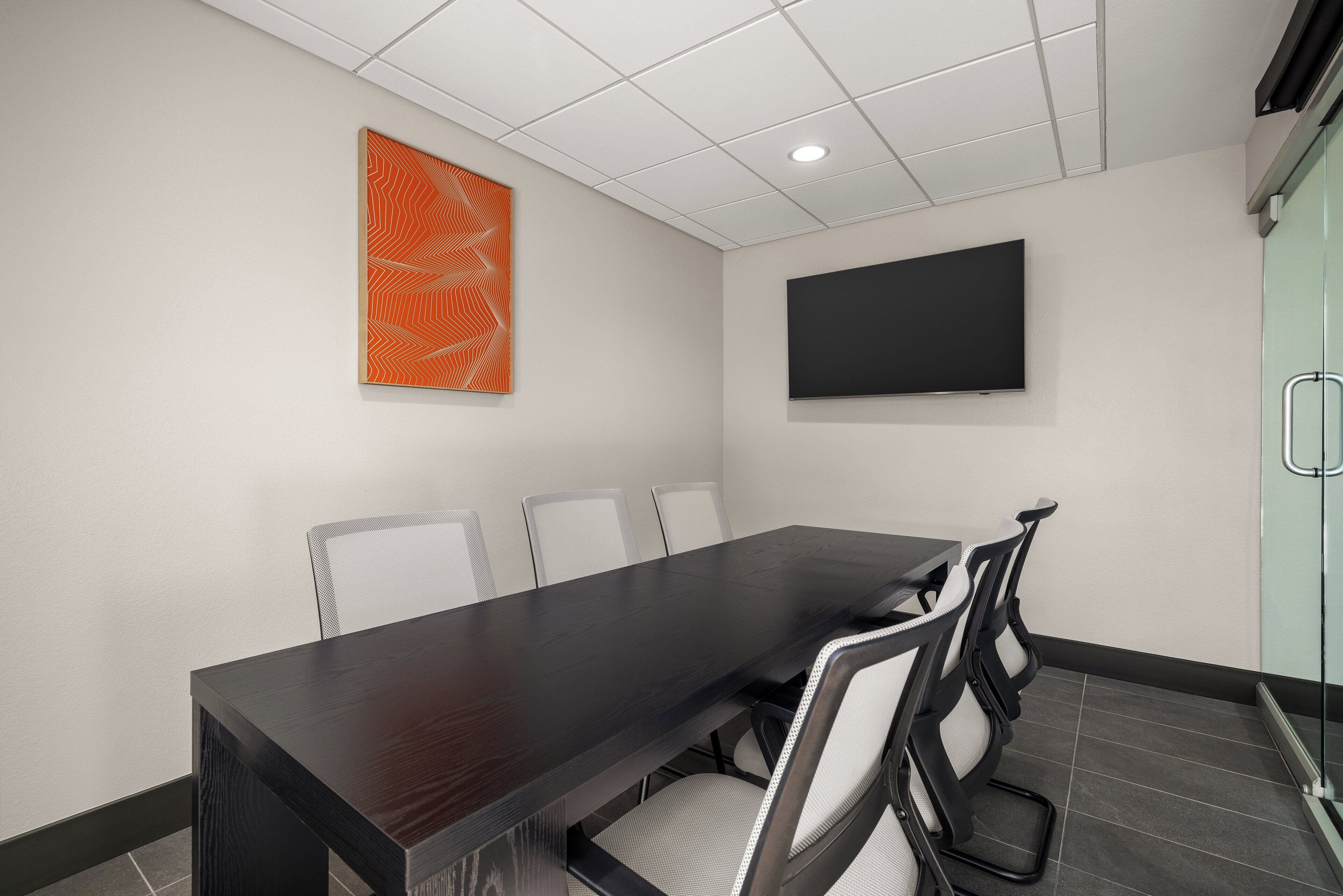 Un salón de conferencias con una larga mesa, sillas blancas, un cuadro abstracto naranja grande y un televisor de pantalla plana en la pared.