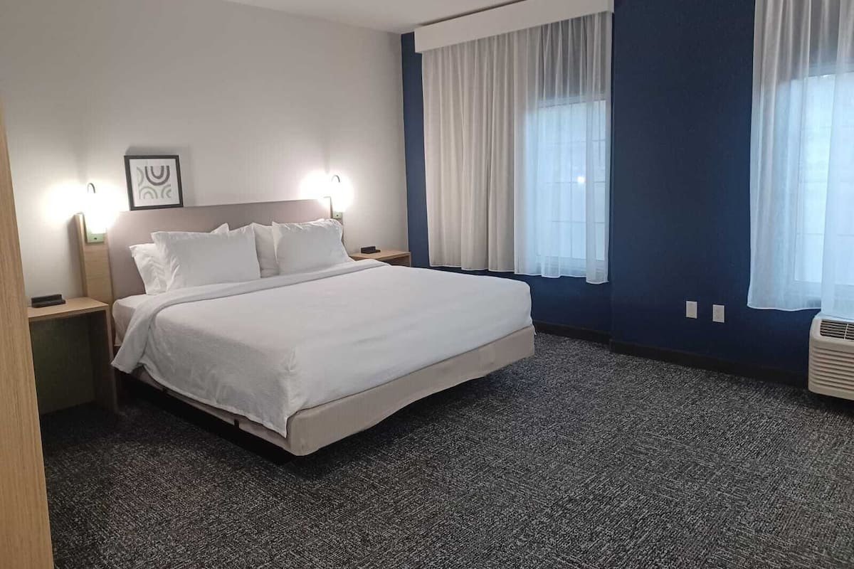 A hotel room with a large bed, two bedside tables, a wall-mounted light, a framed picture, and a window with curtains.