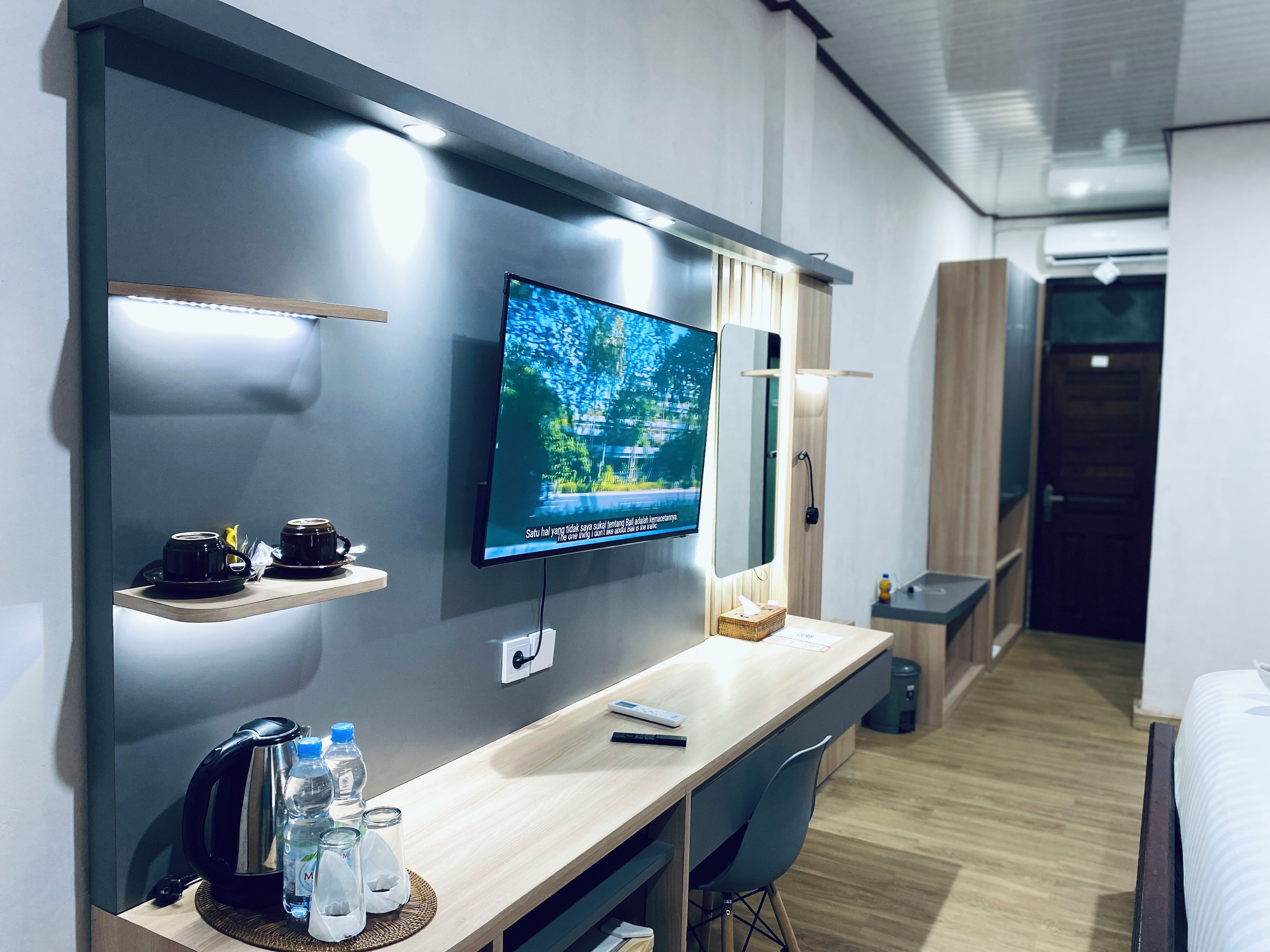 Quarto de hotel moderno com televisão de tela plana na parede, escrivaninha de madeira com chaleira, garrafas de água e controle remoto.