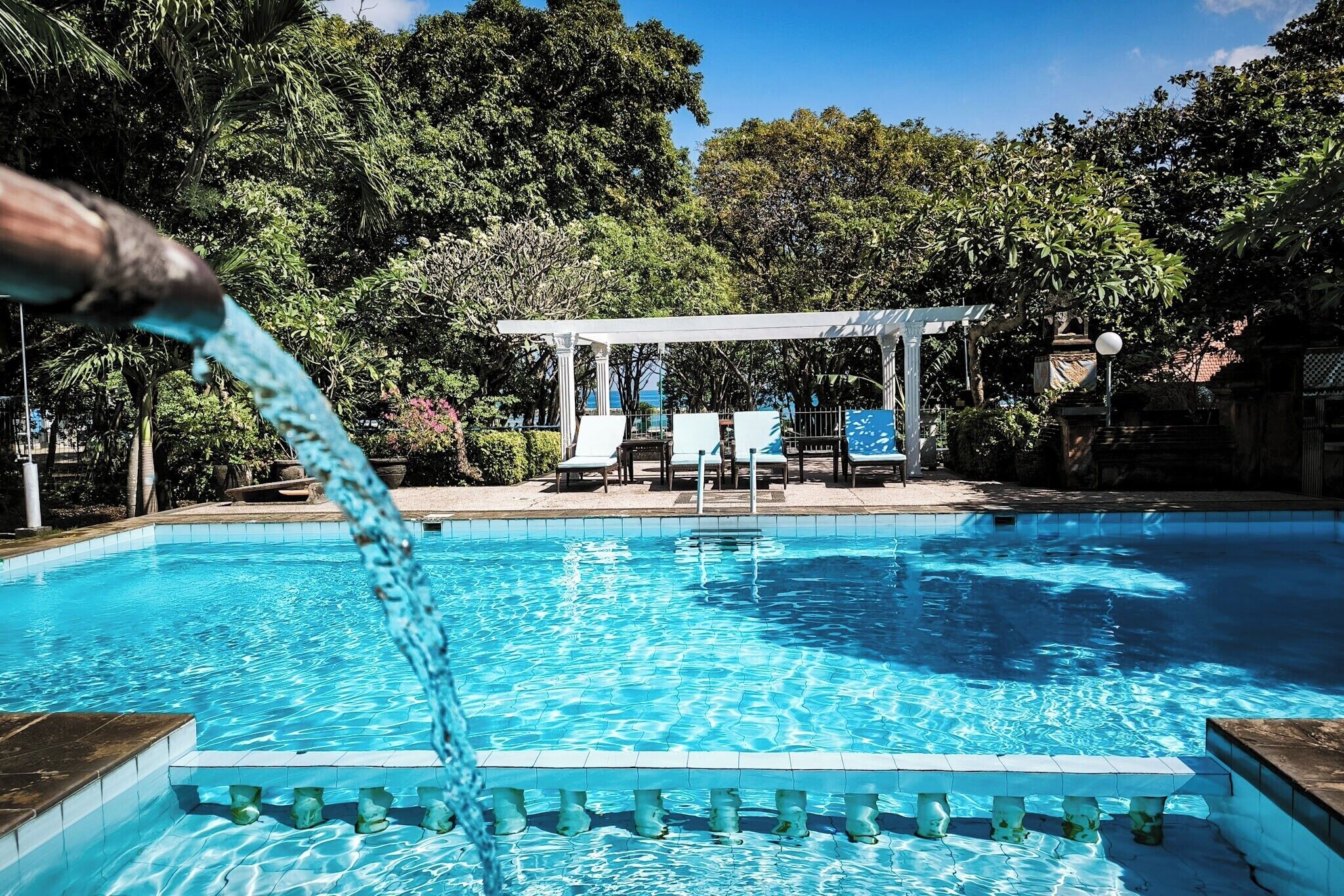 A swimming pool with clear blue water, surrounded by lush greenery and a white pergola with chairs.