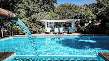 A swimming pool with clear blue water, surrounded by lush greenery and a white pergola with chairs.