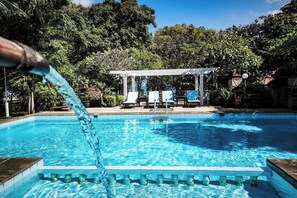 Una piscina con agua azul cristalina, rodeada de abundante vegetación y un porche blanco con sillas.