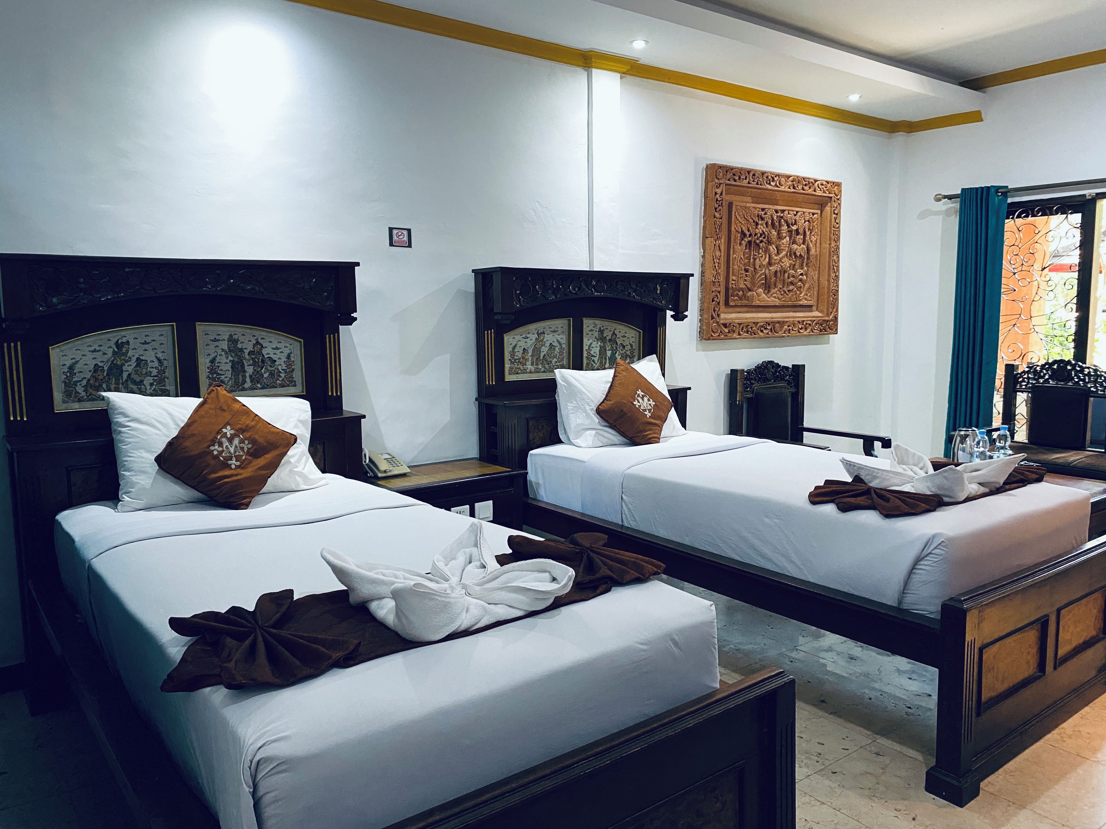 A hotel room with two beds, each with a wooden headboard, white linens, and brown decorative pillows. There is a wall-mounted telephone and a framed artwork above the beds.