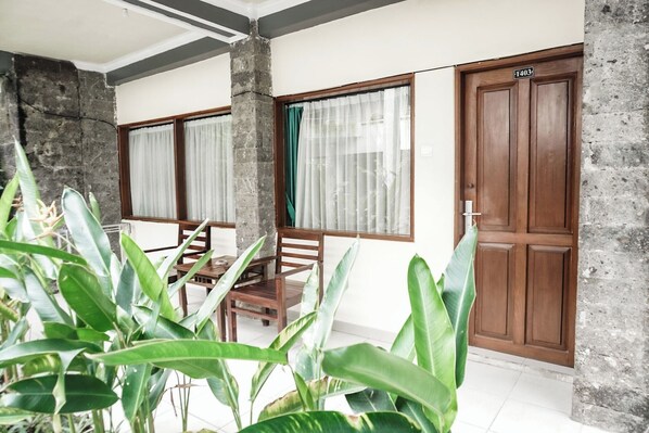 A room with a wooden door, a window with curtains, a table with chairs, and large green plants. - Melasti Kuta Beach Hotel and Spa (Kuta)