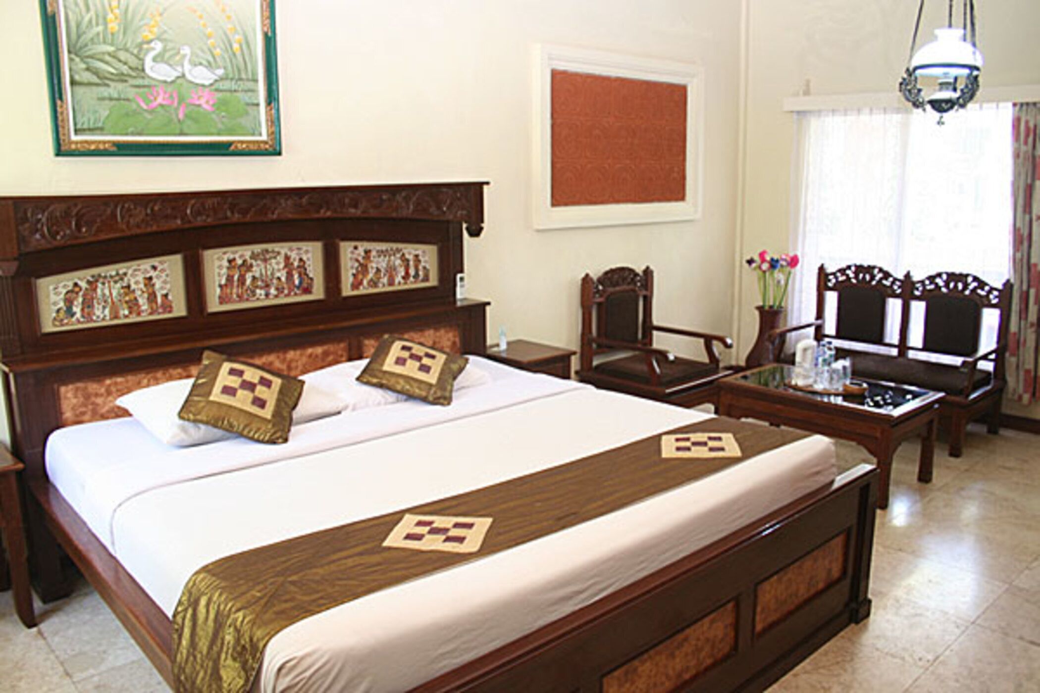 Habitación de hotel con una cama grande, cabecera de madera, dos sillas, una mesita y una ventana con cortinas.