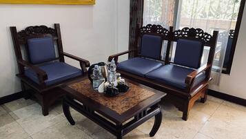 A room with two wooden chairs with blue cushions, a wooden table with a coffee pot, cups, and a tray, and a window with a view of greenery.