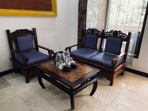 A room with two wooden chairs with blue cushions, a wooden table with a coffee pot, cups, and a tray, and a window with a view of greenery. -  Melasti Kuta Beach Hotel and Spa (Kuta)