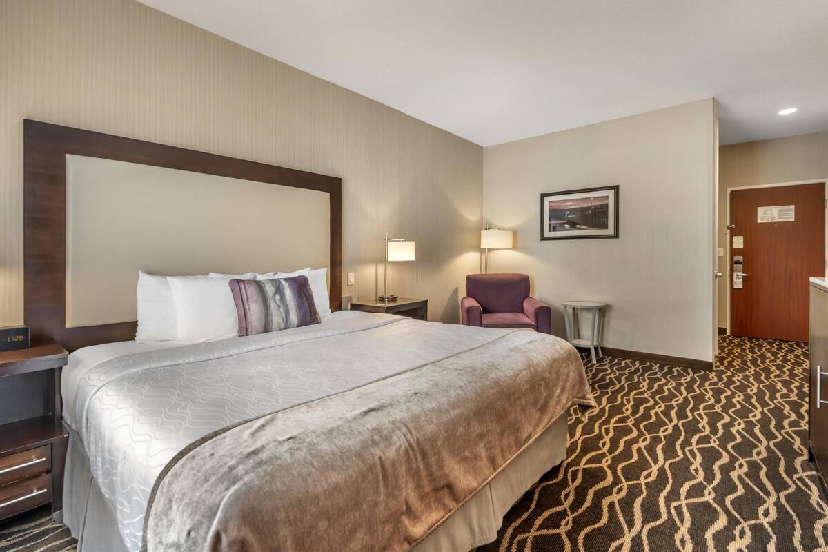 A hotel room with a large bed, a chair, a small table, a lamp, and a door.