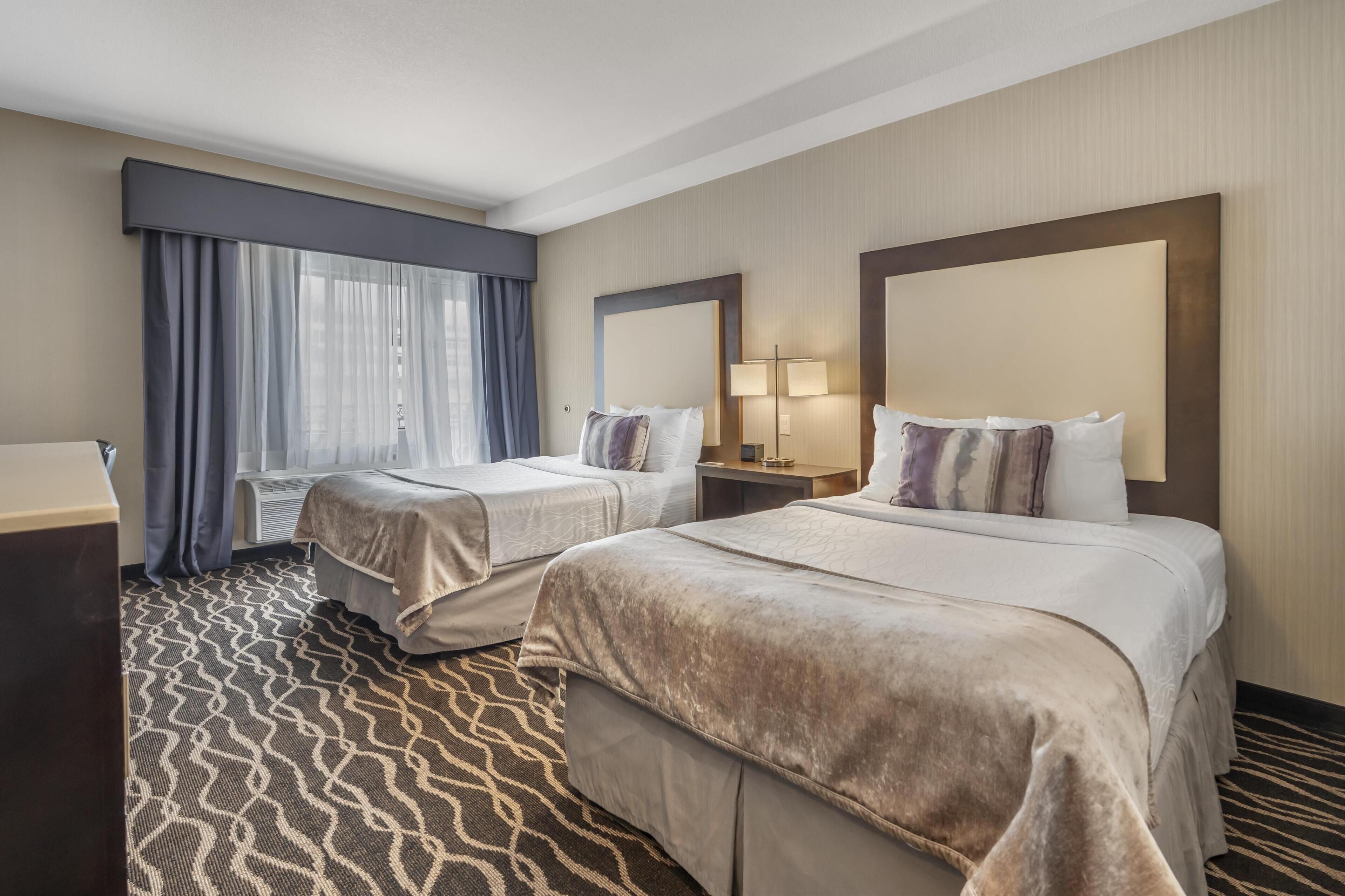 Une chambre d’hôtel avec deux lits, une grande fenêtre avec des rideaux, une moquette à motifs et une table de chevet avec une lampe.