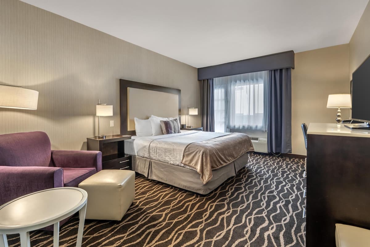 A hotel room with a bed, a desk, a chair, a small table, and a large window with curtains.